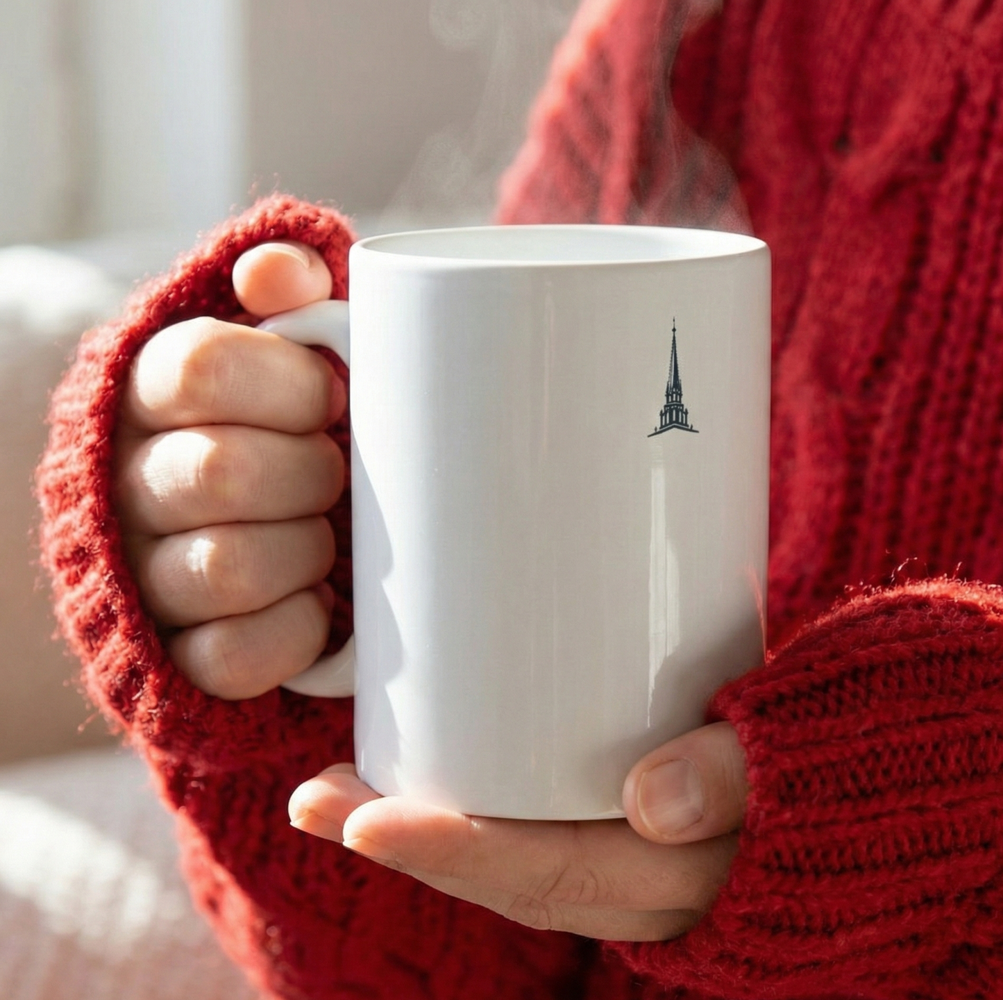 Gros plan sur un mug blanc avec un logo flèche de Notre Dame de Paris, tenu par des mains vêtues d'un pull en laine rouge vif, ambiance lumineuse. Le cadeau parfait pour les noces de plomb et pour marquer 14 ans de mariage en France et en Pologne, un moment chaleureux et mémorable.